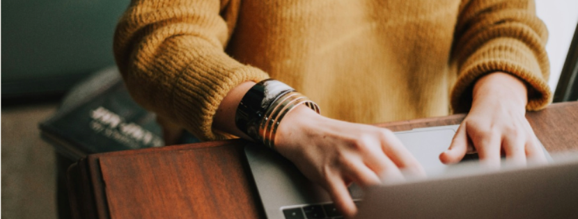 A person wearing a yellow sweater and multiple bracelets is typing on a laptop. The image is taken from an angle above the person's left shoulder, showing the hands on the keyboard and part of the laptop screen. The background is blurred, with a hint of a book and possibly a desk or table.